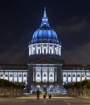 Nighttime view of a blue-lit domed civic building with people walking on the plaza.
