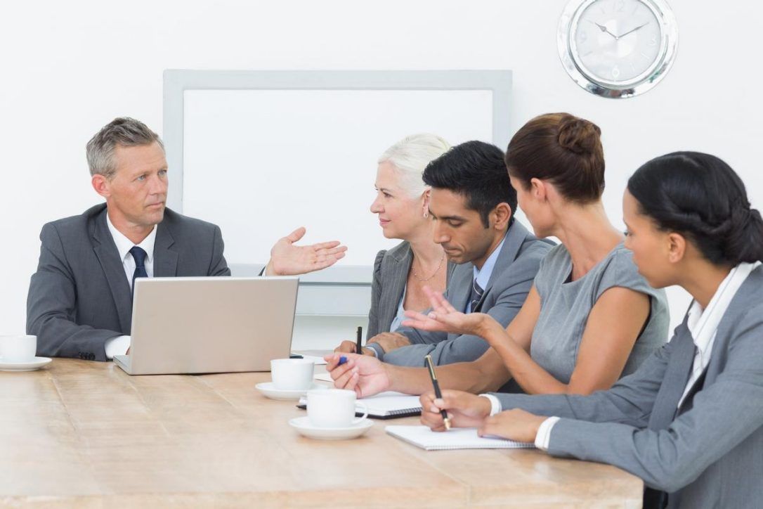 A businessman leads a conversation in the boardroom while typing on his laptop