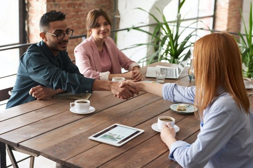 A professional woman shakes hands with a man sitting across the table from her to confirm a deal