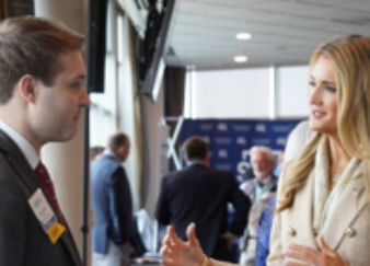 Two professionals talking at a networking event, with tables and people in the background.
