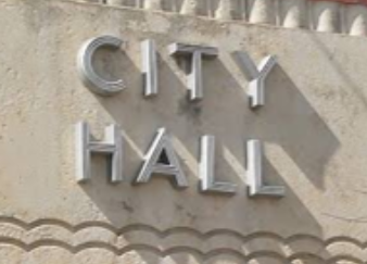 “CITY HALL” sign on a beige building facade with a brick wall above it.