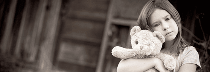 Young girl holding a teddy bear, leaning against a wooden wall in a sepia-toned photo