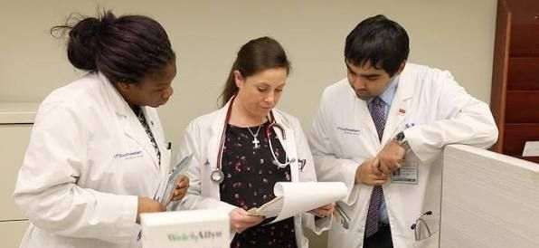 Three clinicians in white coats reviewing a patient chart together in a hospital hallway