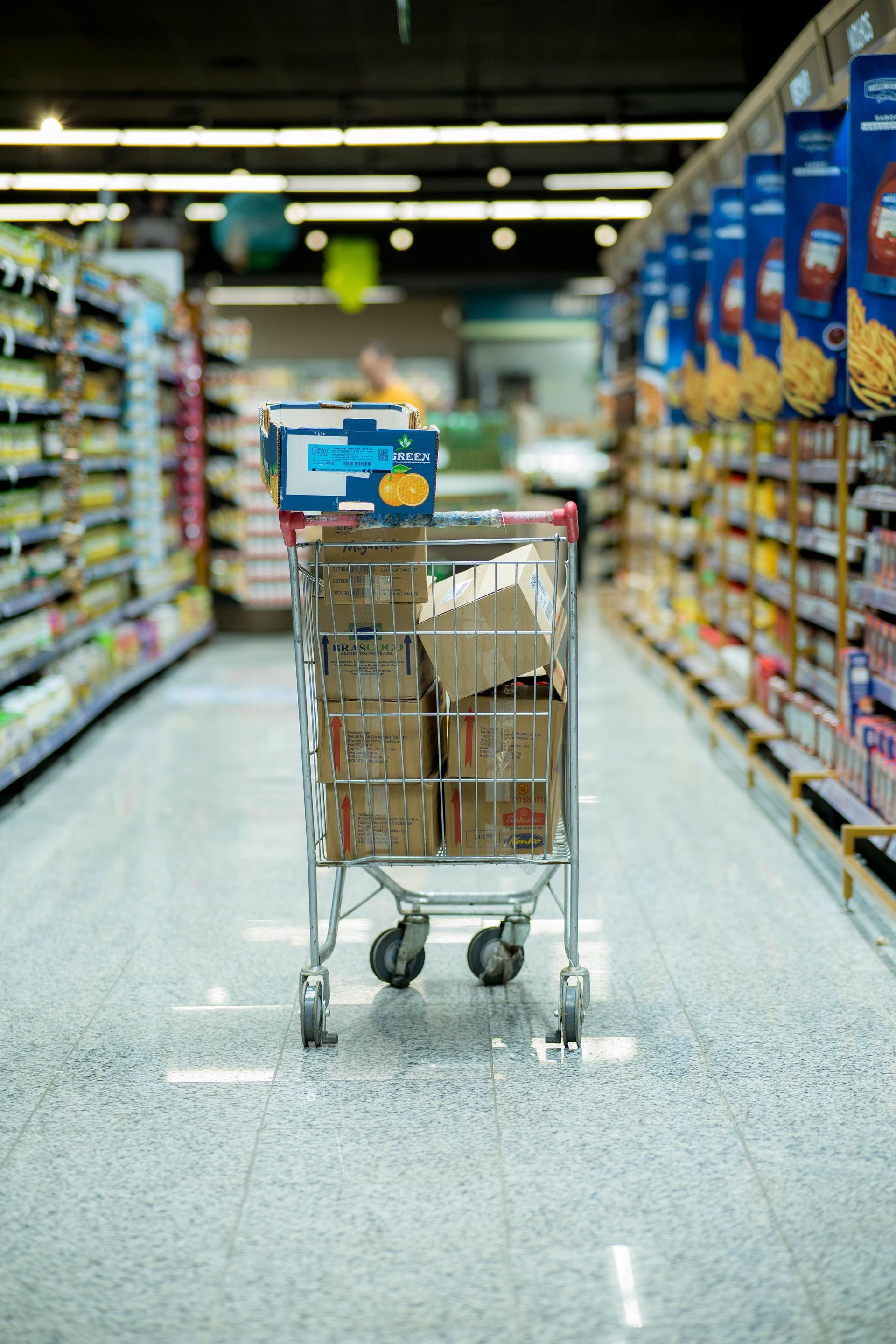 Shopping cart with groceries in a supermarket aisle, boxes stacked inside, shelves blurred in the background