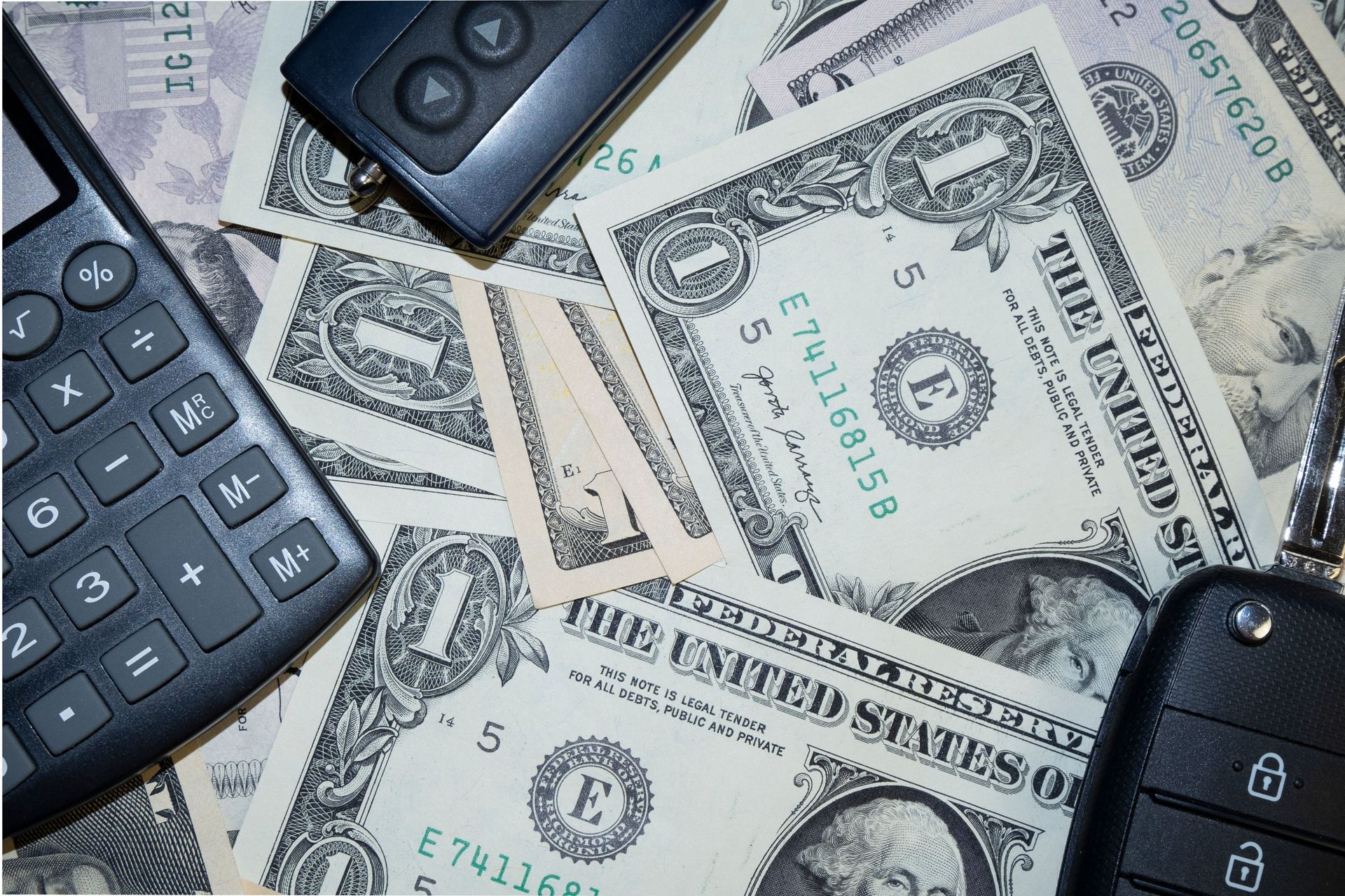 Calculator and black devices scattered over U.S. dollar bills on a table.