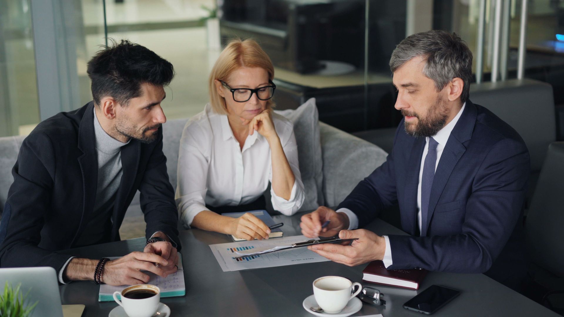 Three coworkers in suits review a document at a conference table with coffee cups in a meeting room.