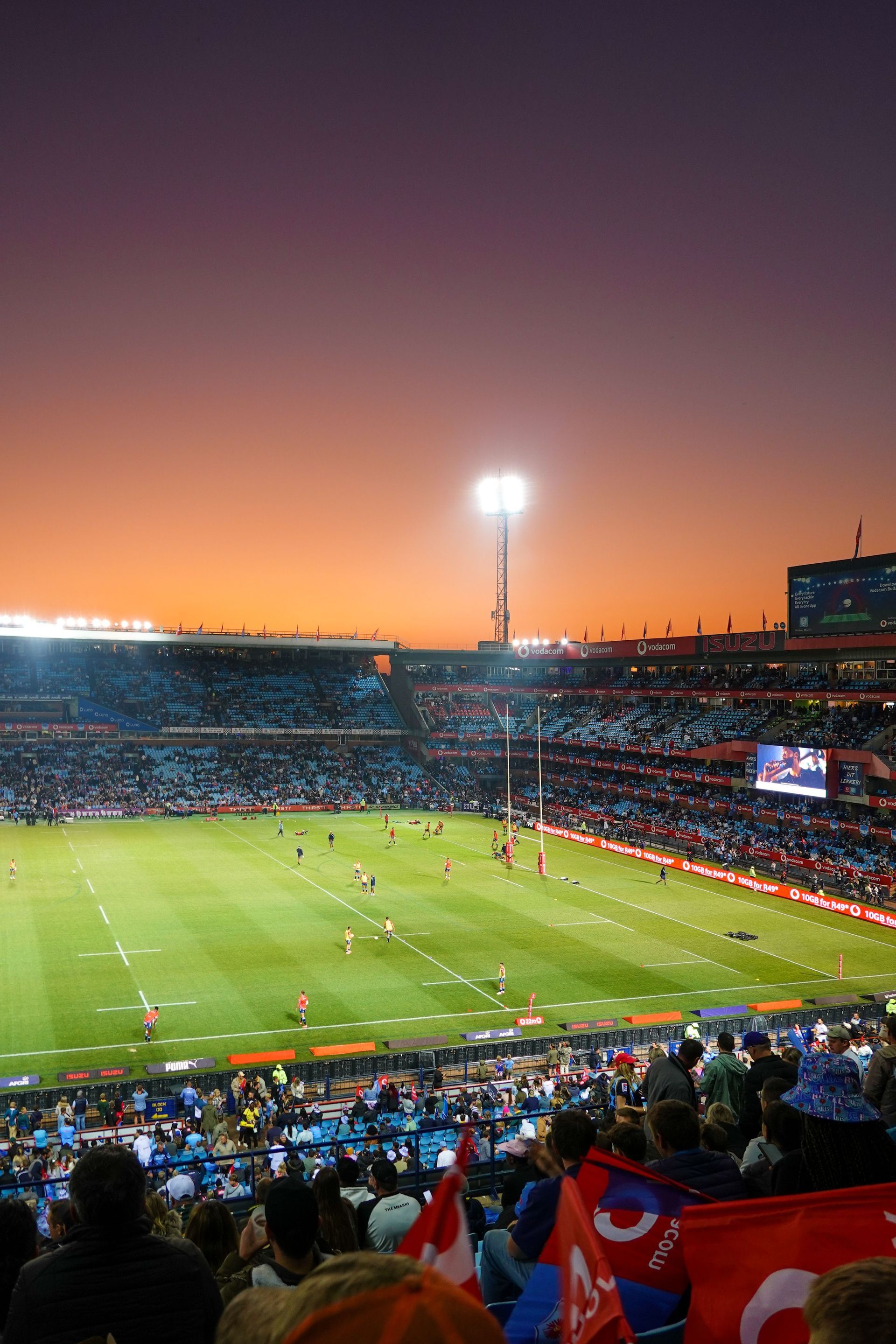 Soccer match at a packed stadium at sunset, with bright floodlights and fans in the stands.