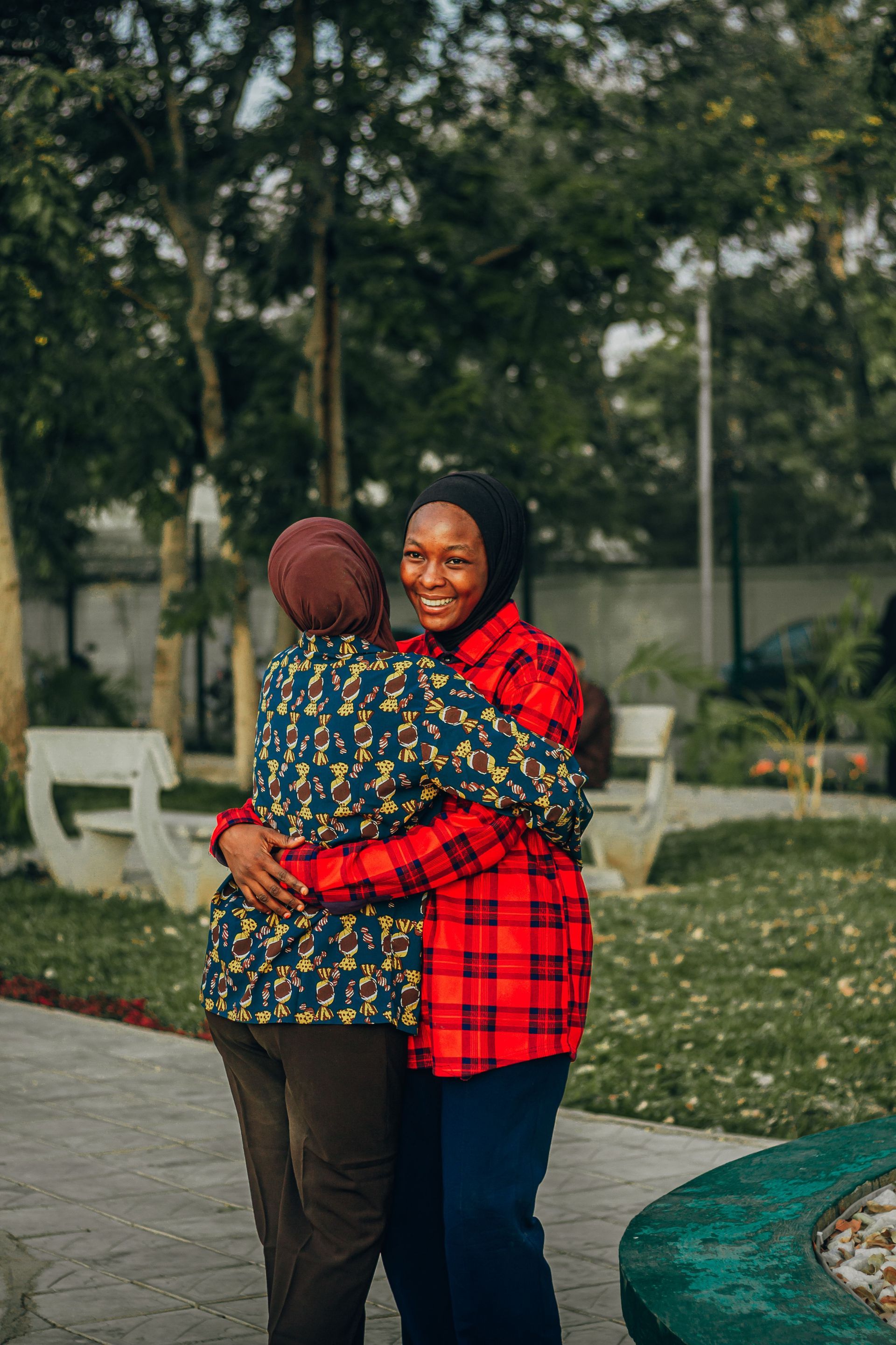 Two people hugging in a park, one in a red plaid shirt and both smiling near a fountain.