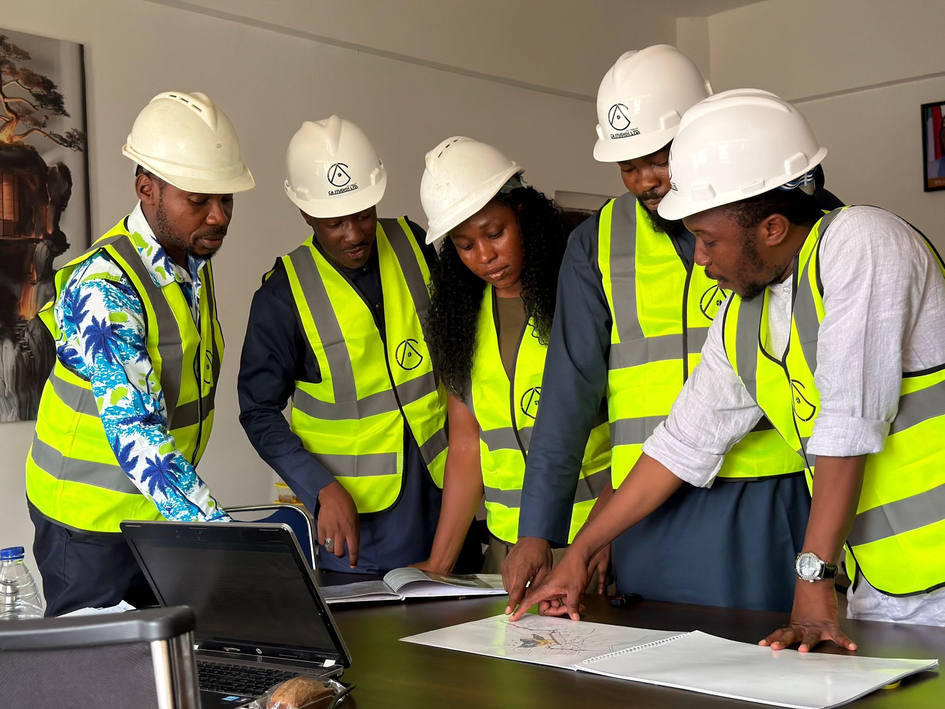 Five workers in hard hats and safety vests review plans around a laptop at a table
