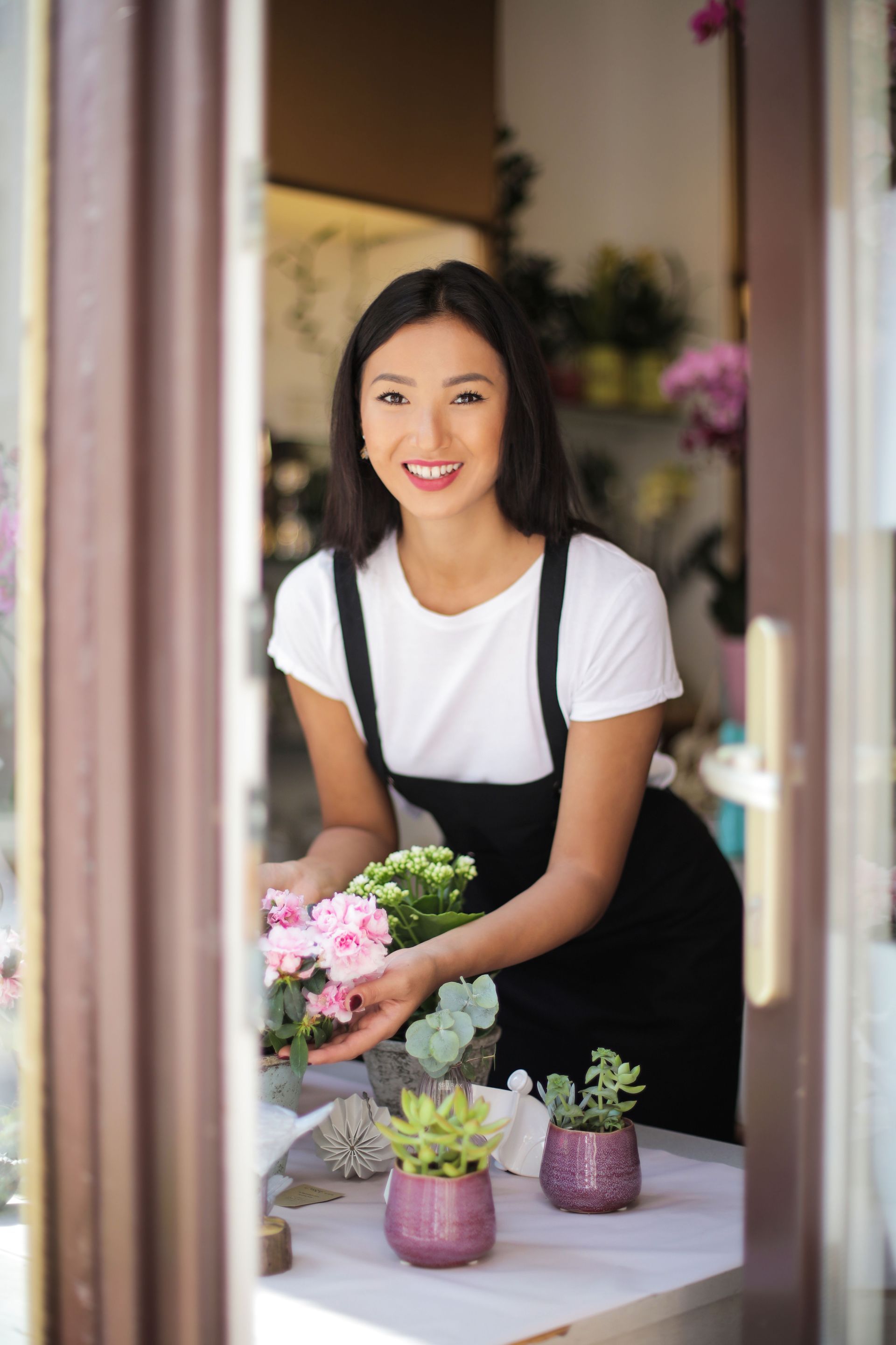 Smiling florist arranging pink flowers at a table with small potted succulents by a doorway