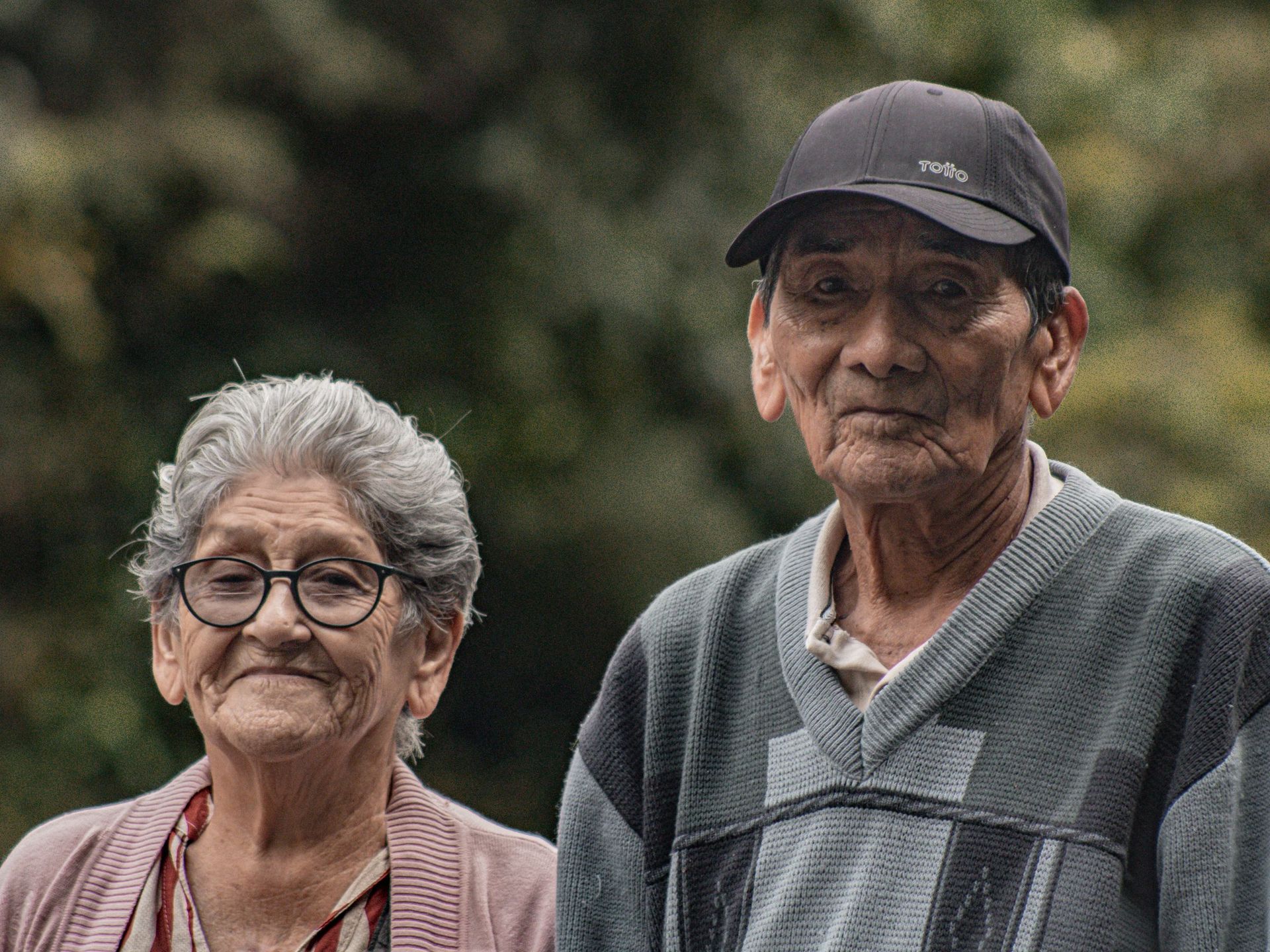 Two elderly people standing outdoors, wearing glasses and a cap, with a soft green background