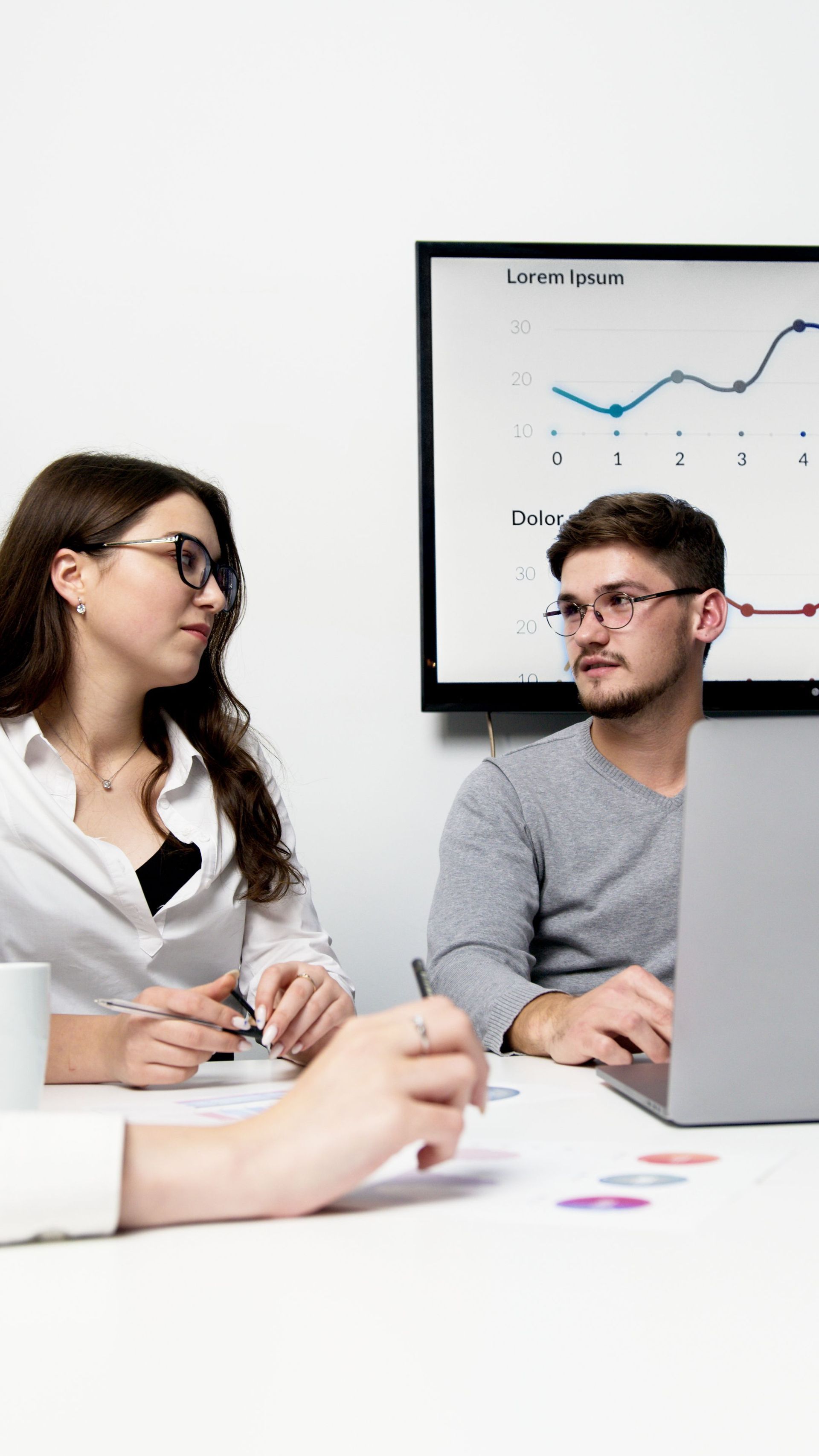 Three coworkers in a meeting, talking at a table with a laptop and chart on a wall display