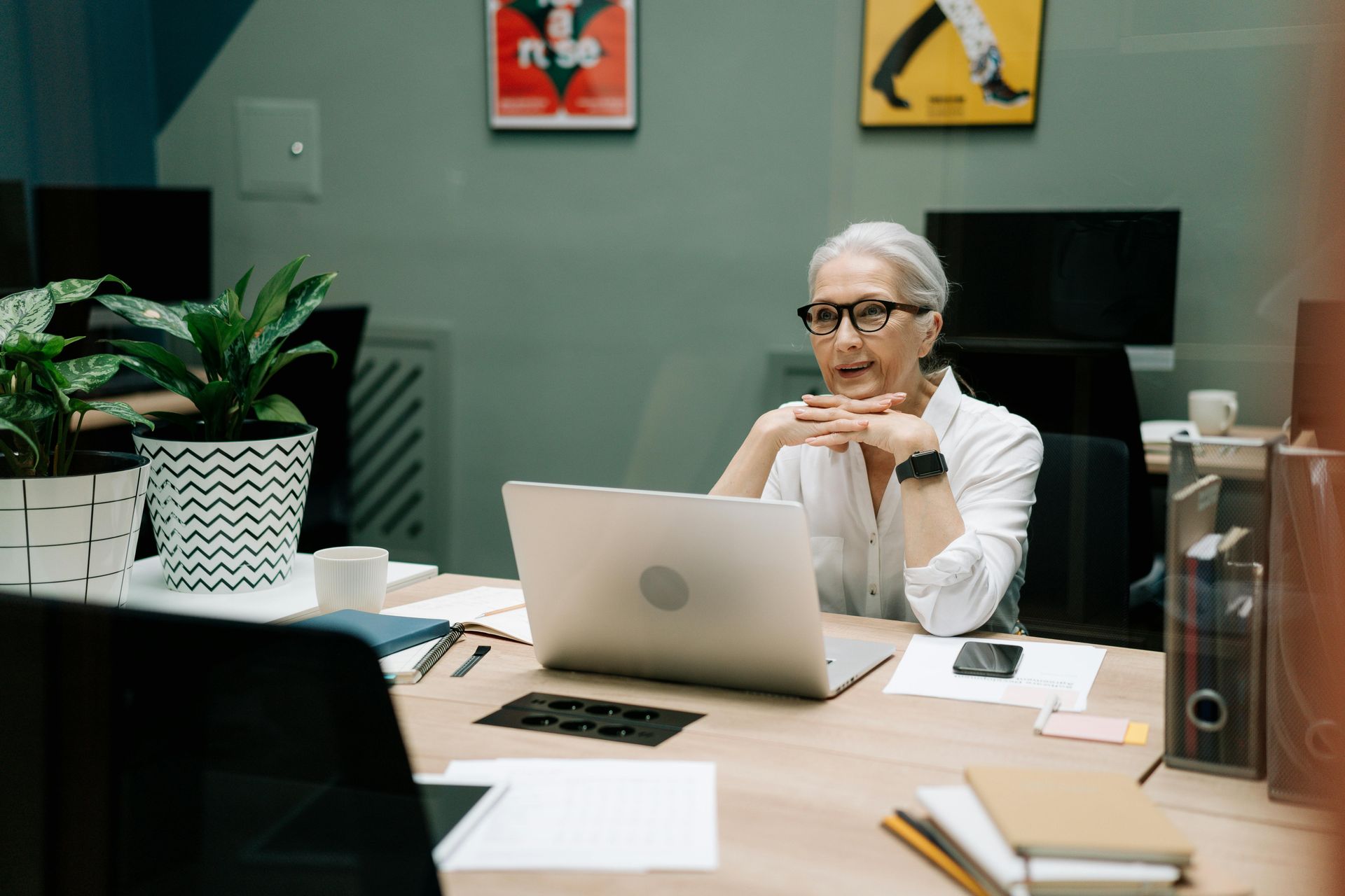 Woman in glasses sits at a desk in front of a laptop, hands clasped, looking at the screen.