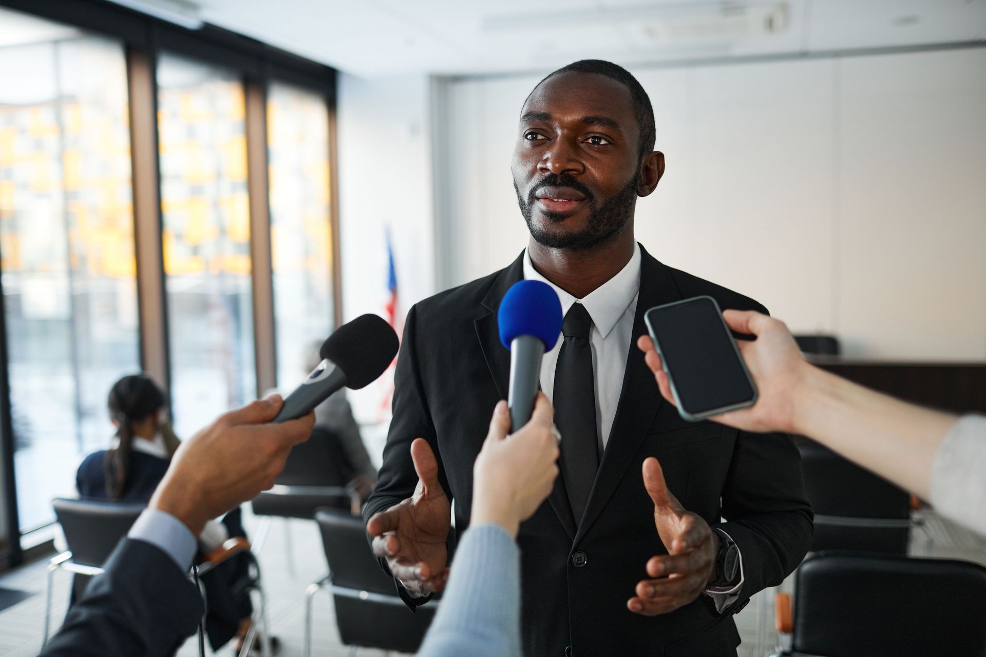 Man speaking at a press conference, surrounded by microphones and smartphones in a bright room