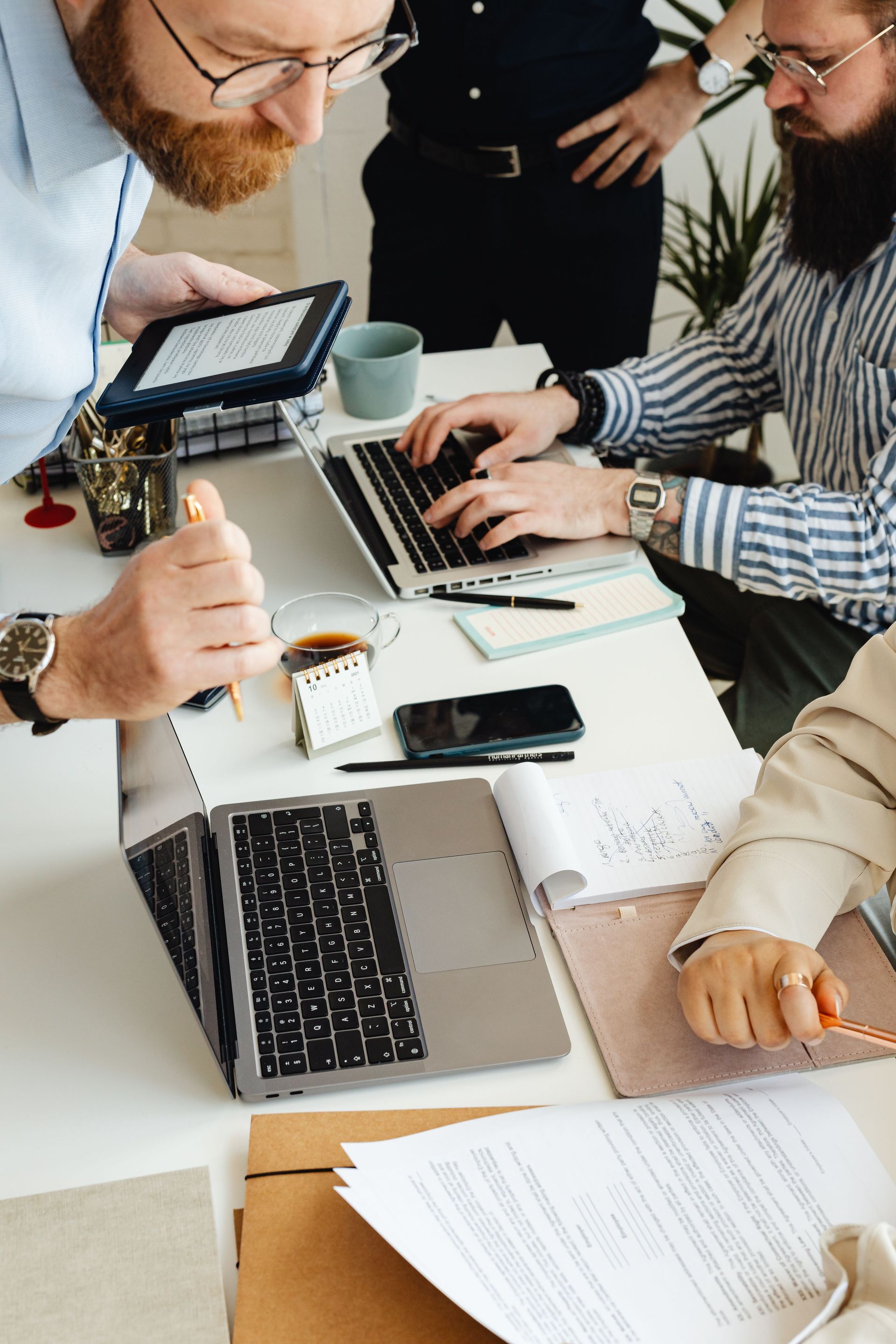 People collaborating around laptops at a white office table with papers, coffee, and a tablet.