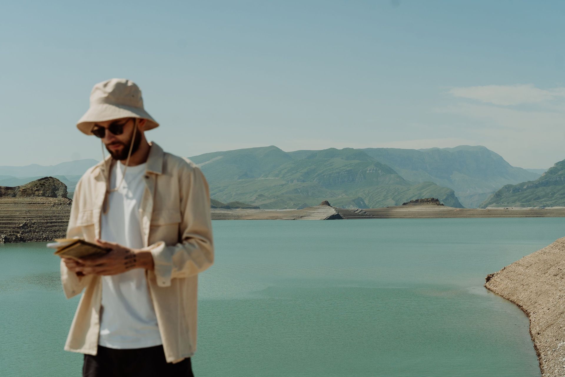 Man reading a book beside a turquoise lake with mountains in the background