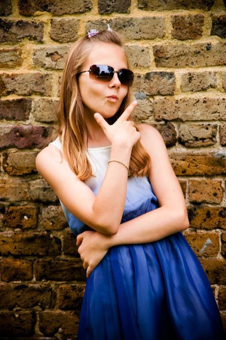 A young girl wearing sunglasses and a blue dress is standing in front of a brick wall.