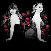 Two women are sitting back to back in front of a bush of red roses.