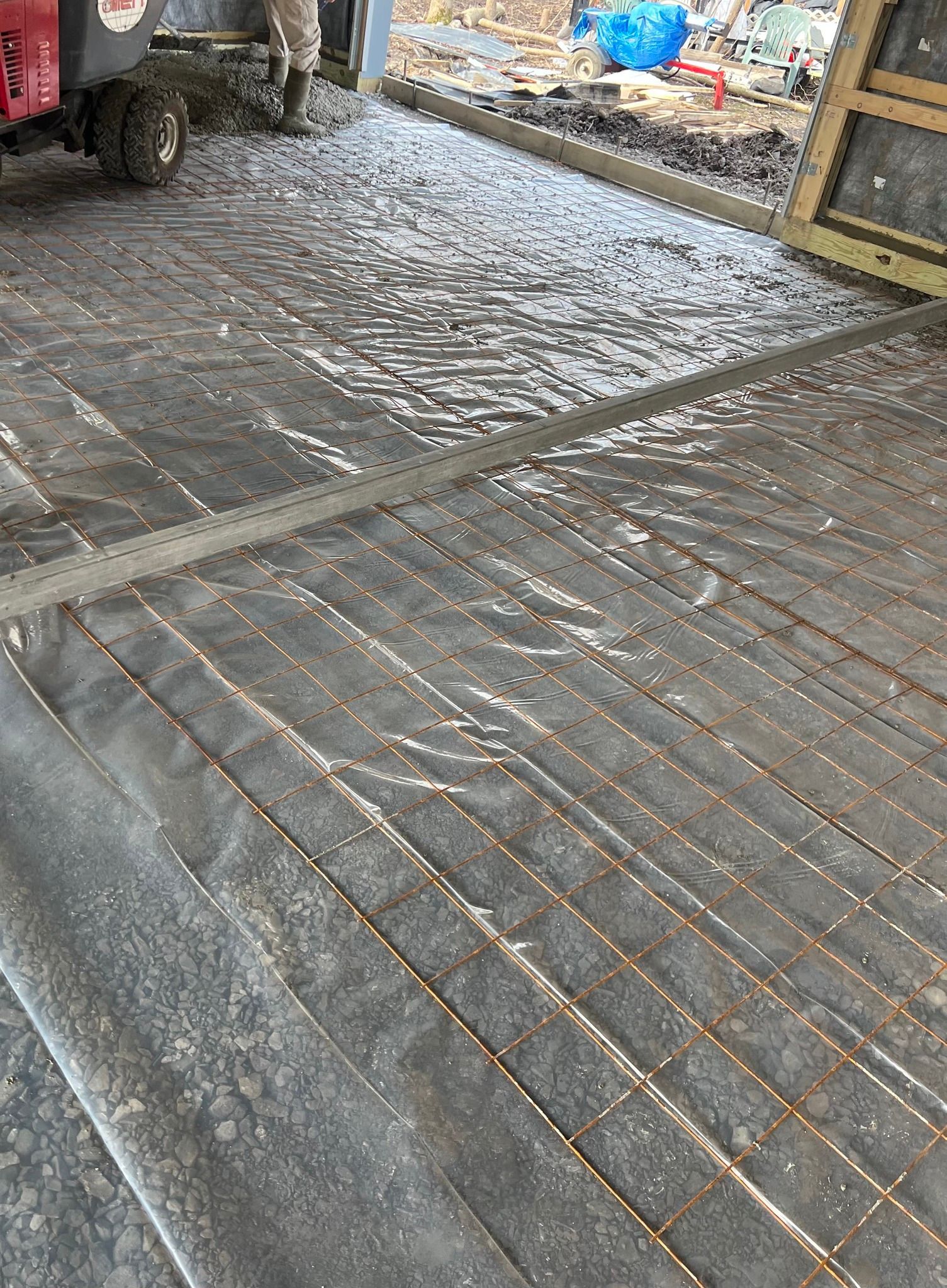 A wire mesh grid laid over a plastic vapor barrier on a construction site, with a piece of heavy equipment in the corner.
