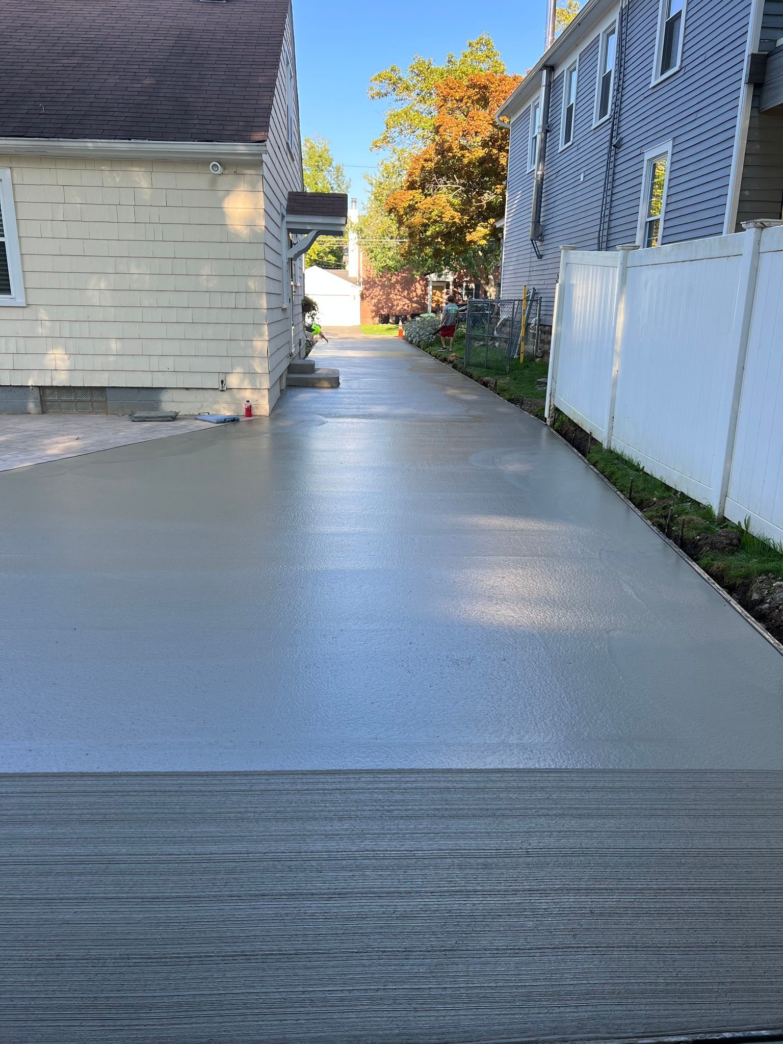 A newly paved gray concrete driveway between a yellow house and a blue house with a white fence.