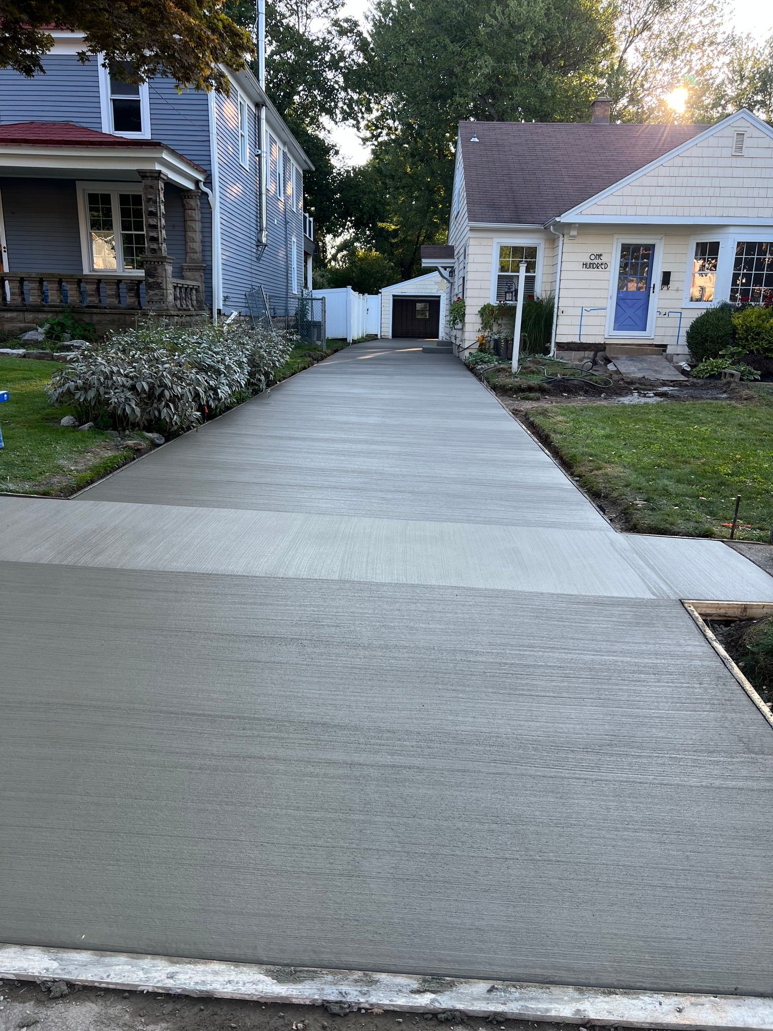 A newly poured, gray concrete driveway extends between two houses toward a detached garage at sunset.