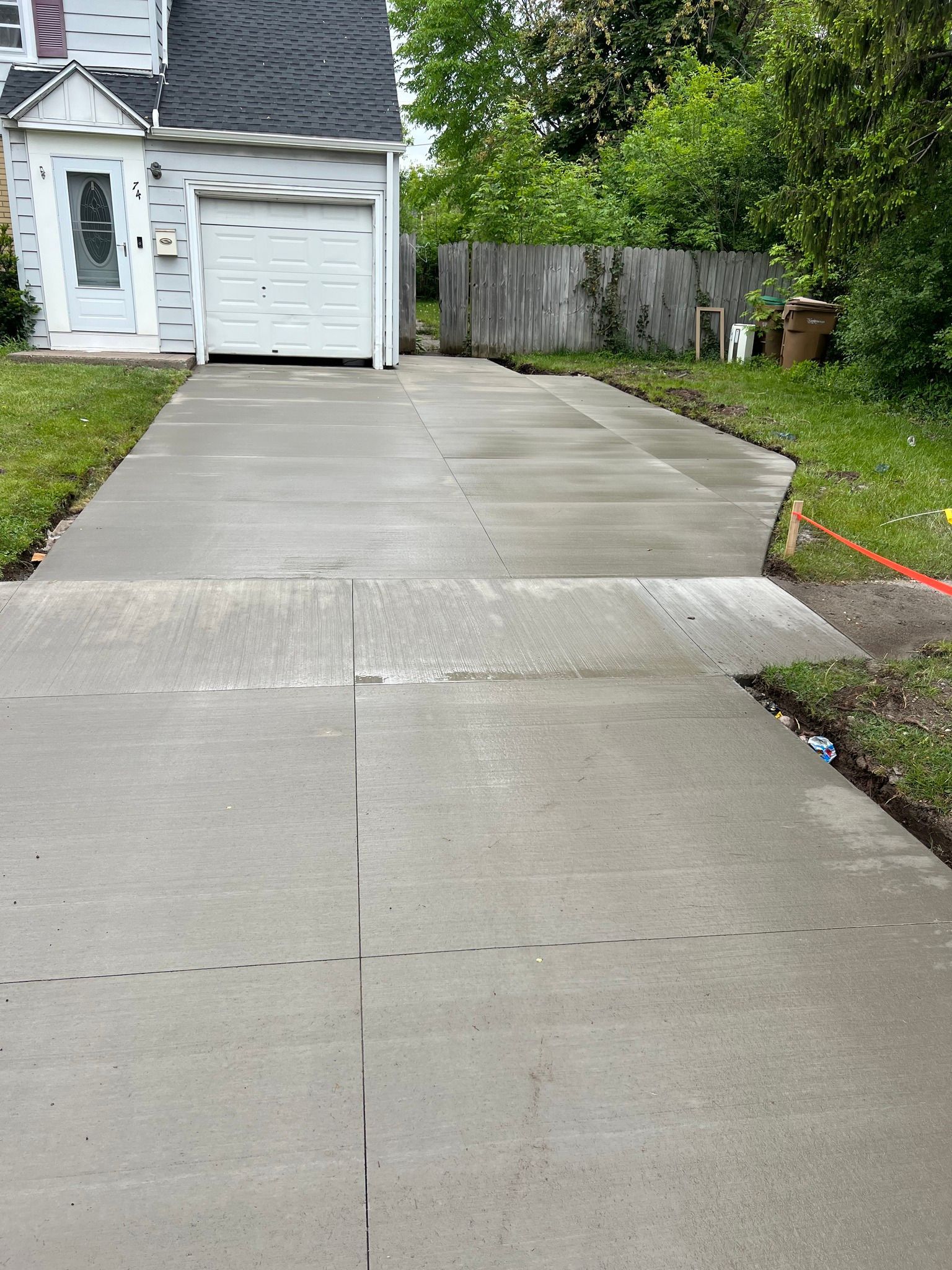 A recently paved grey concrete driveway leading to a white house with a garage, bordered by green grass and a wooden fence.