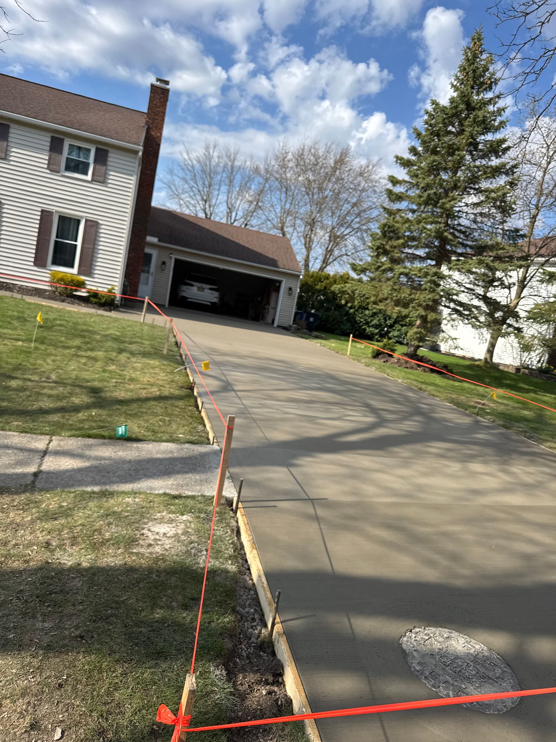 A residential driveway under construction, bordered by orange caution tape and wood stakes, leading to a house and garage.