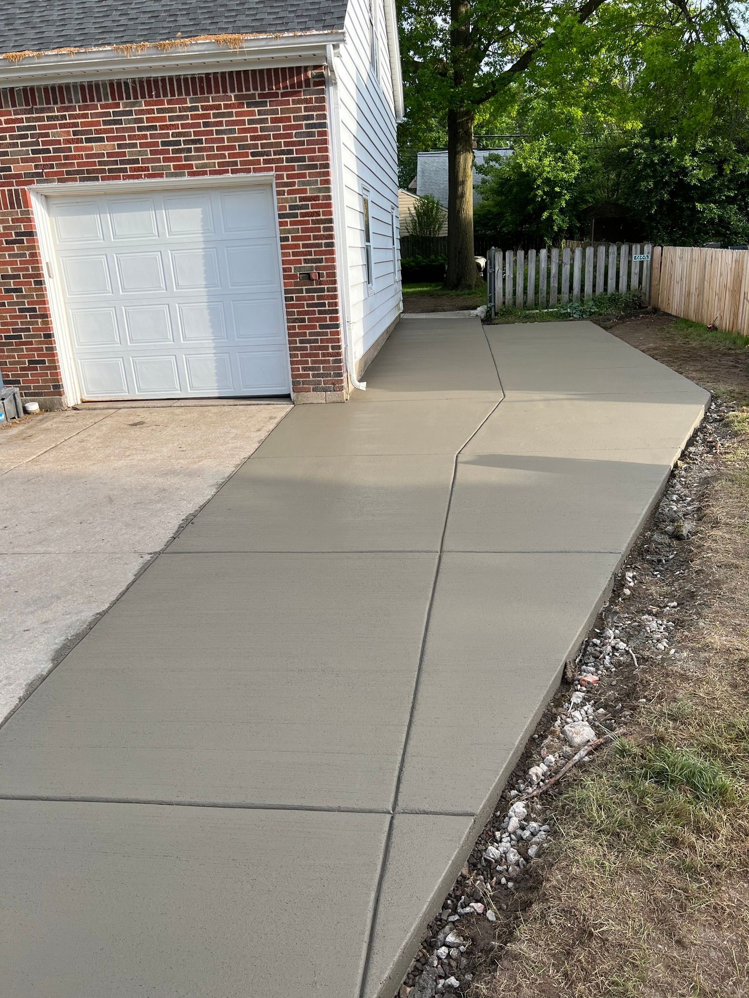 Newly poured gray concrete sidewalk leading alongside a brick residential garage toward a wooden fence and trees.