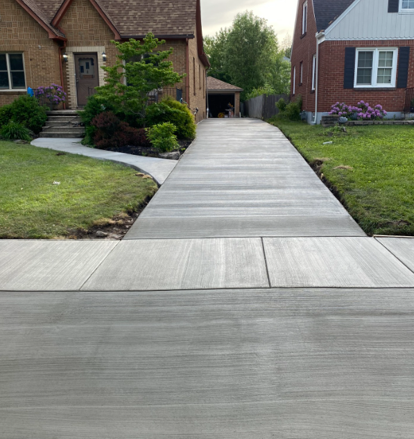 A freshly poured concrete driveway leads from the sidewalk toward a brick house and garage on a suburban street.