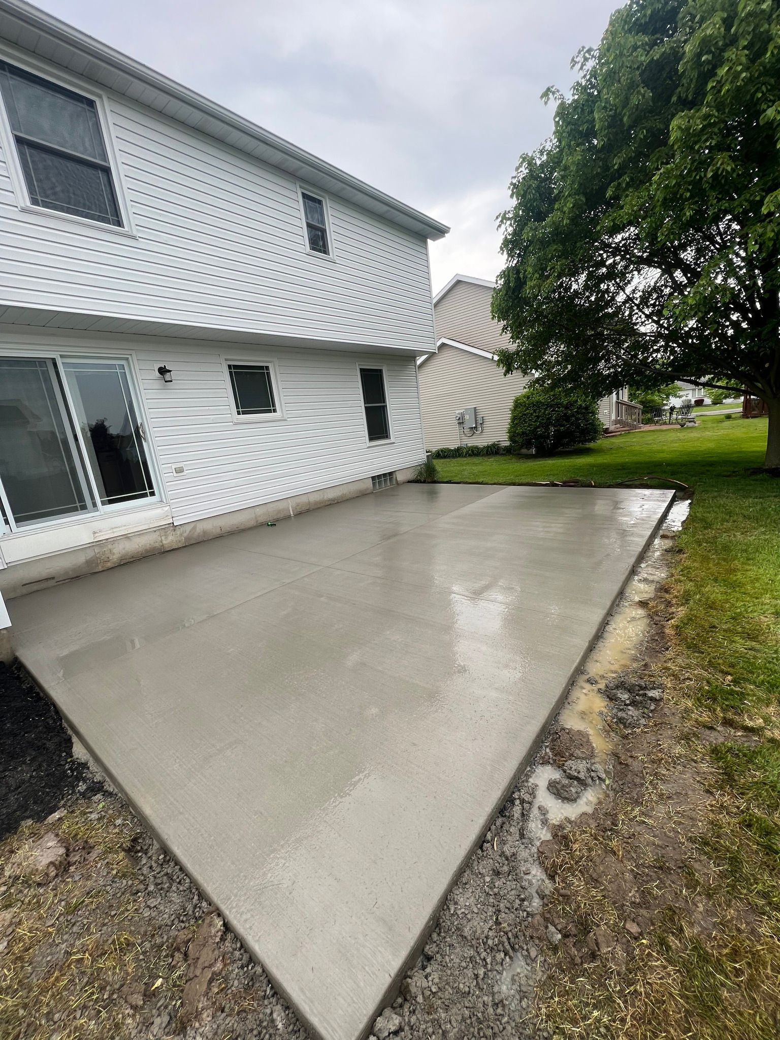 A freshly poured concrete patio attached to the back of a two-story white house with siding on a cloudy day.