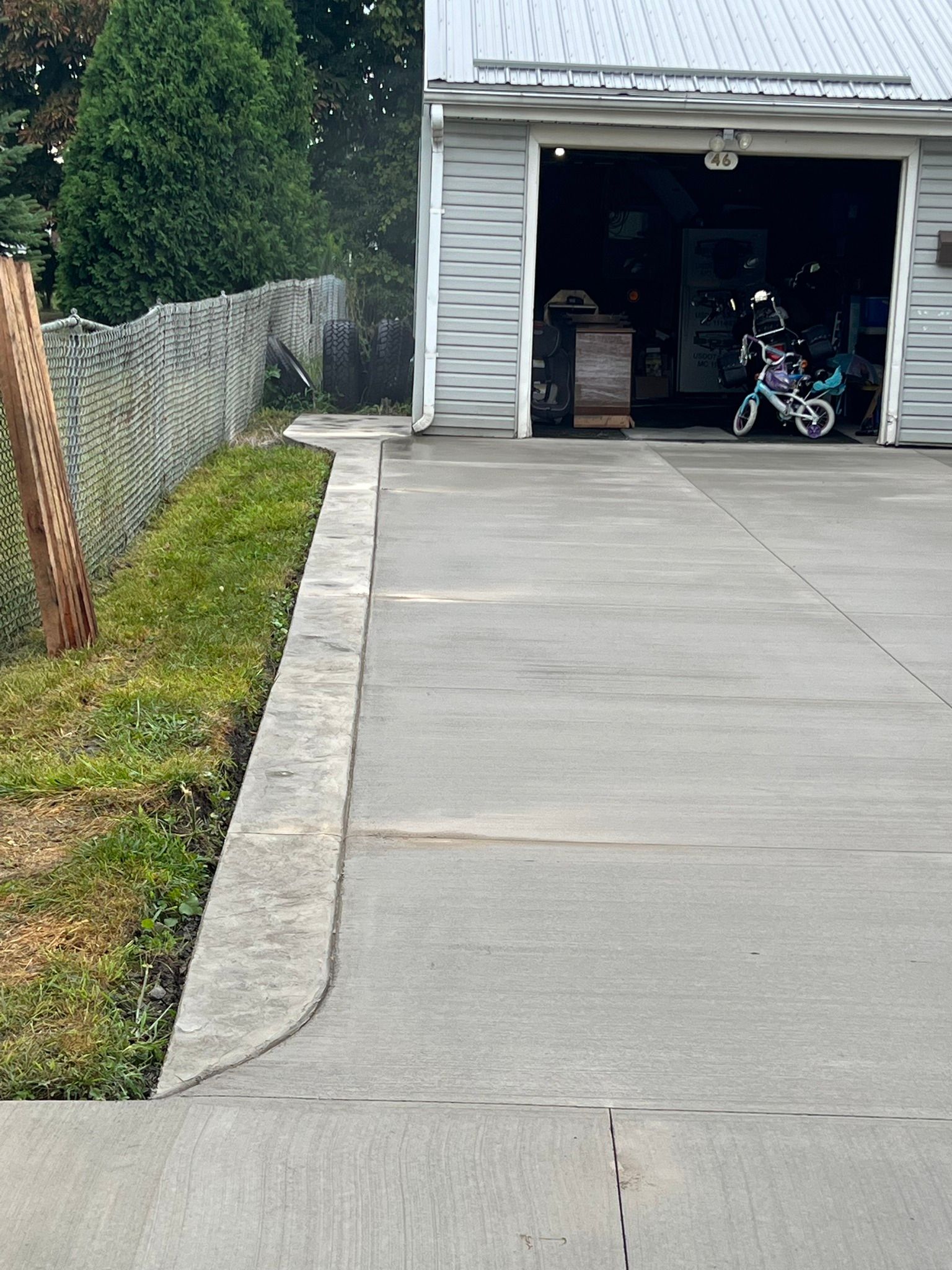 A concrete driveway leads to a garage with a metal roof, alongside a grassy patch and a wire fence.