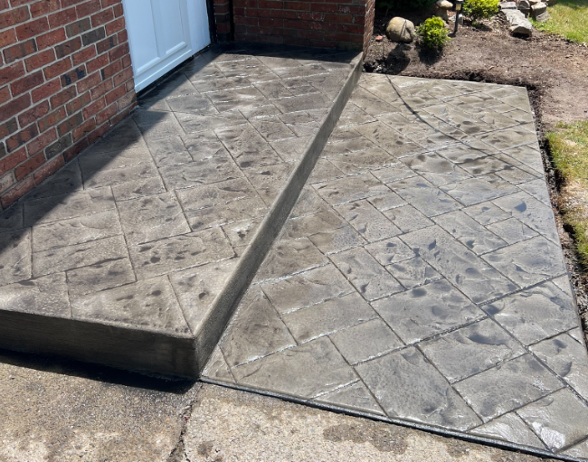A newly poured, dark gray stamped concrete walkway and step featuring a diamond pattern next to a brick house.