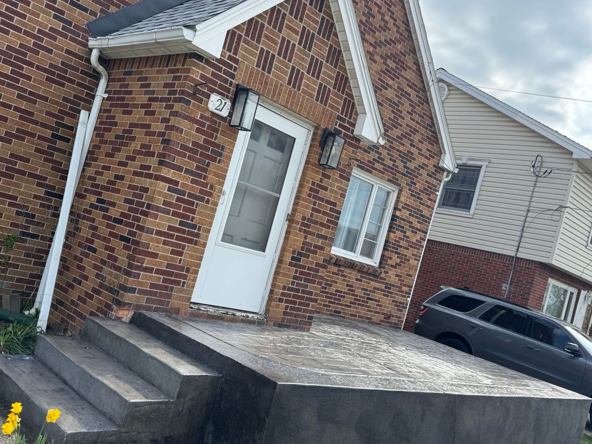 A brick house entrance with a front porch, stone steps, a white door, and a gray SUV parked on the side.