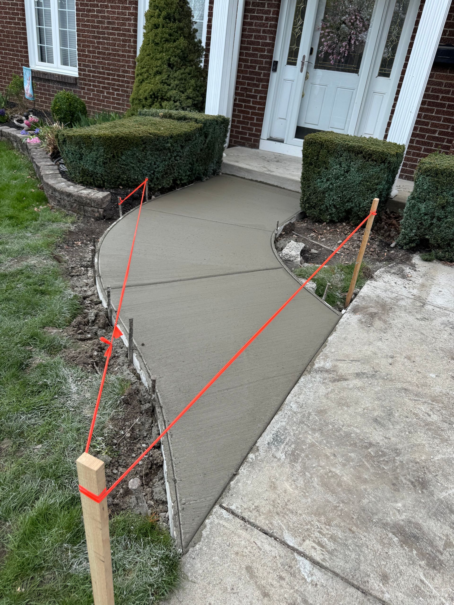 A freshly poured concrete sidewalk path leading to a brick house front entrance, marked off by wooden stakes and orange cord.