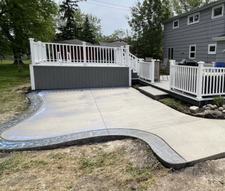 A gray raised deck with white railings sits beside a house, overlooking a new concrete patio with a decorative border.