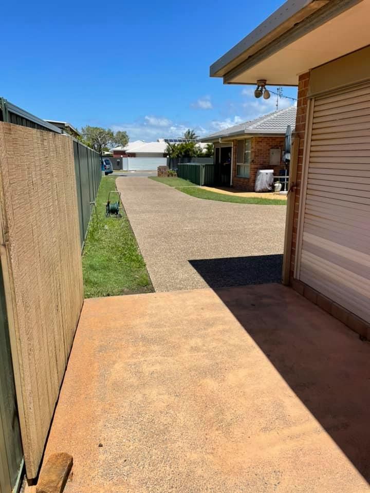 Narrow concrete driveway between a wooden fence and a brick building, leading to other houses under a blue sky.