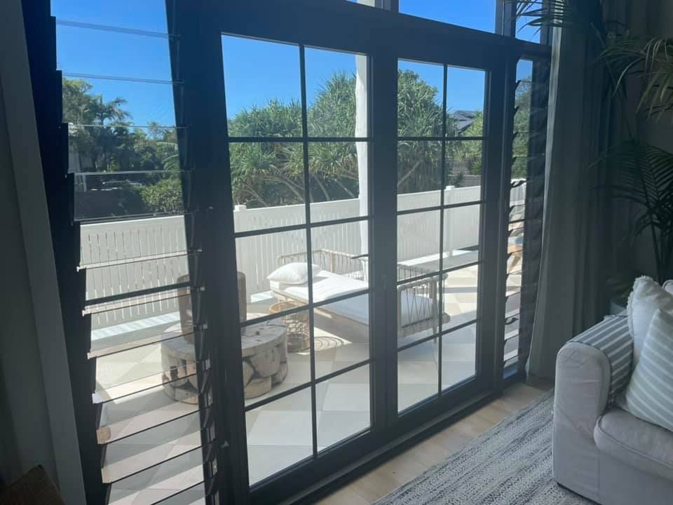 Black-framed glass doors open to outdoor patio. Patio includes seating, white fence, and greenery. Interior features sofa and rug.
