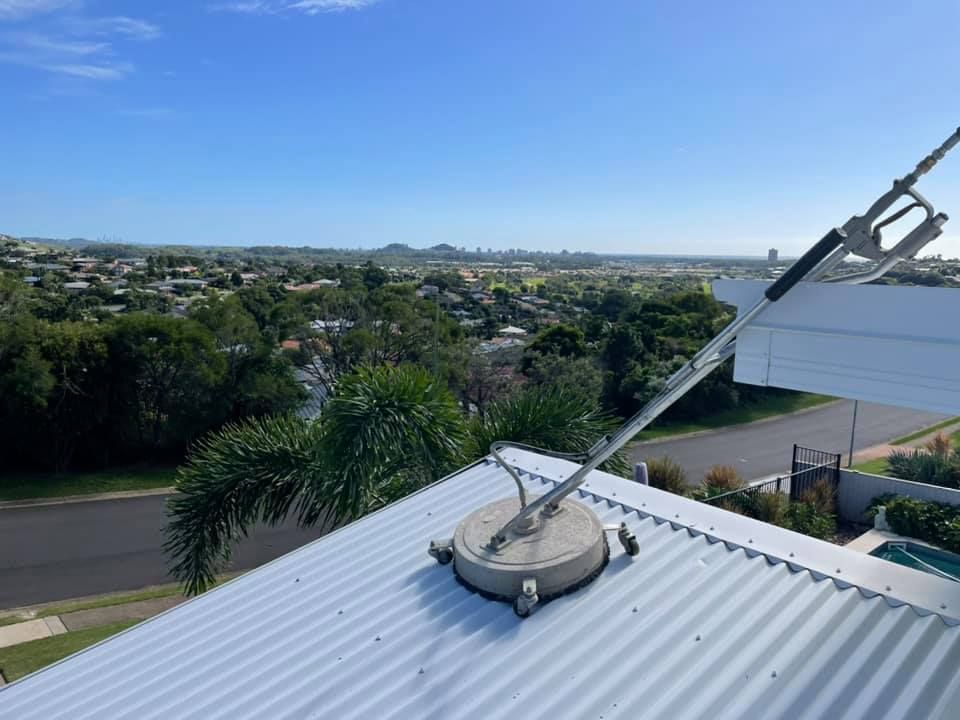 Pressure washer cleaning a metal roof with a suburban landscape in the background under a blue sky.