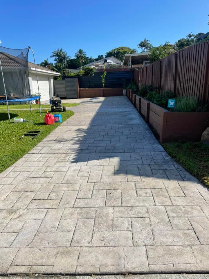 Concrete driveway leading to a house, flanked by raised garden beds and a wooden fence, on a sunny day.