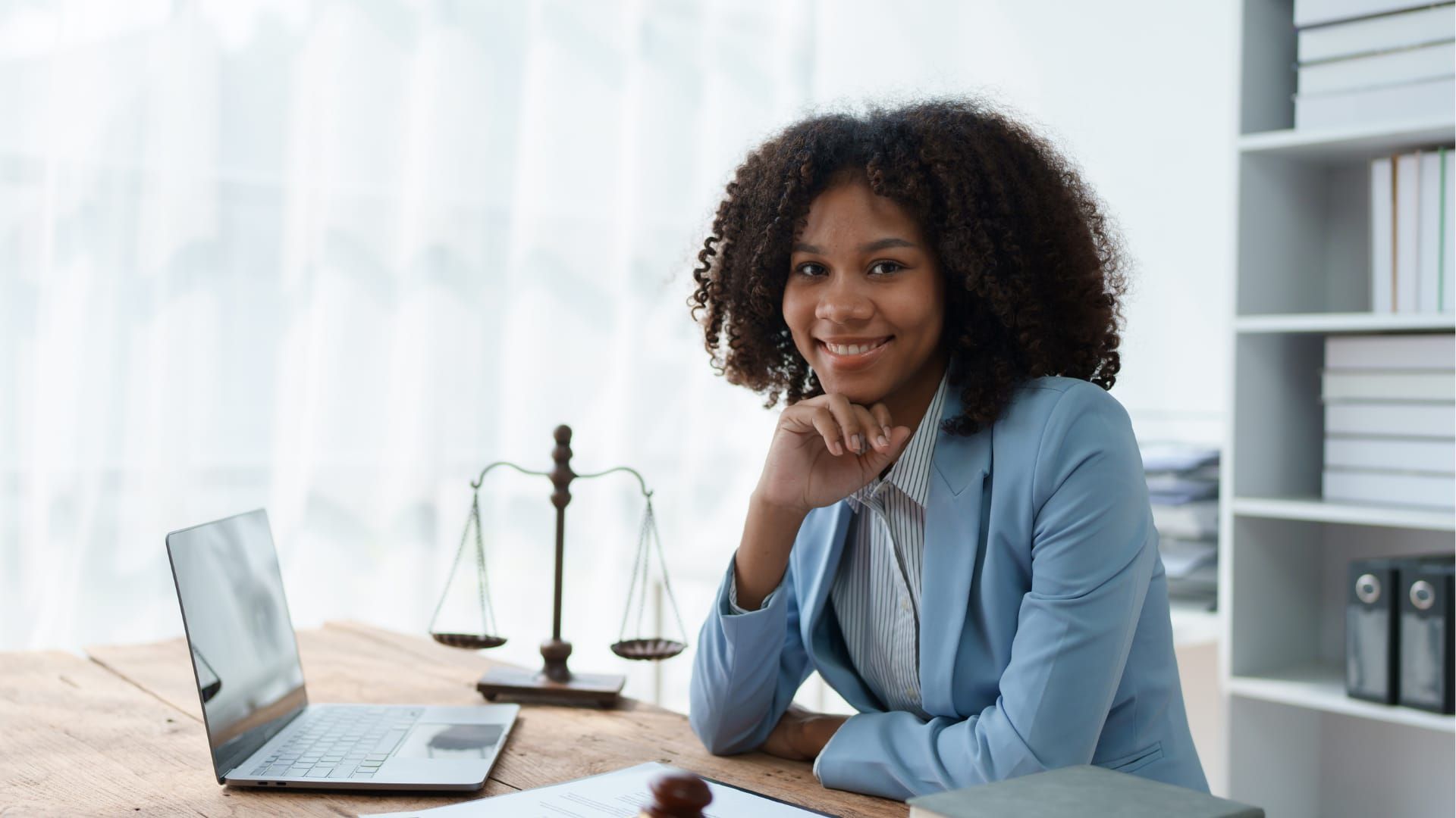 Black woman lawyer smiling, at desk with laptop, scales of justice, and books. Wearing a blue blazer.