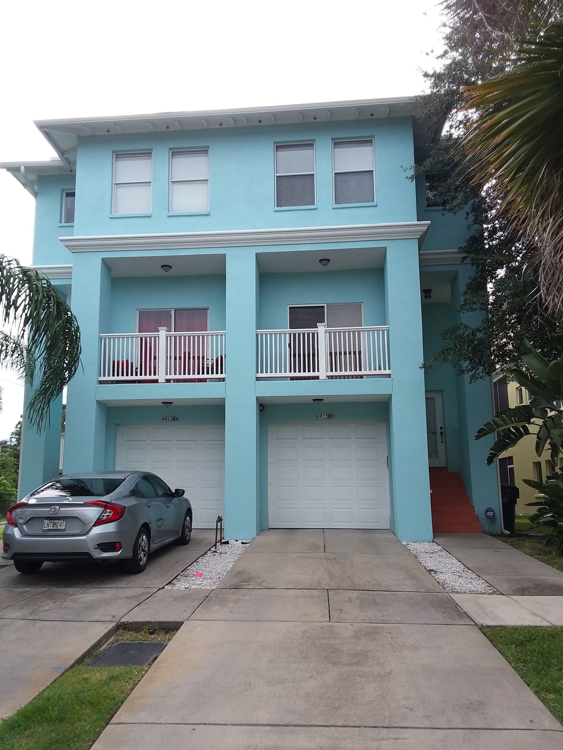 A light blue multi-story building with two garage doors, balconies, and a car in the driveway.