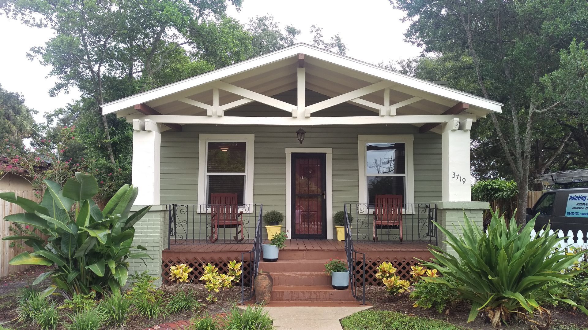 Green bungalow with porch, plants, and white accents; front view.