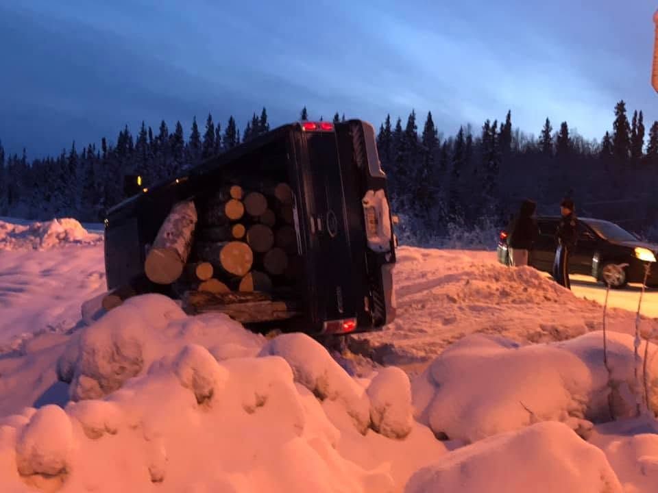 Overturned log truck in snowy roadside ditch at dusk, with another vehicle nearby and dark pine forest behind.