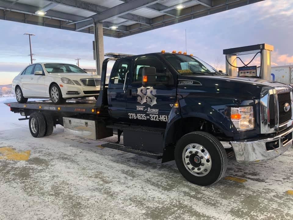 Tow truck carrying a white sedan under a snowy covered lot at dusk