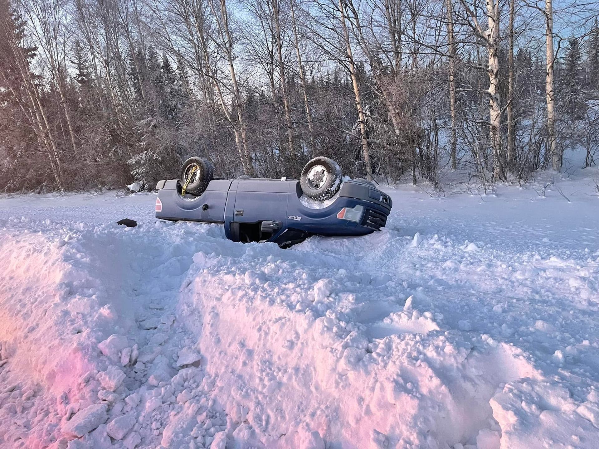 Overturned blue car in deep snow near a snowy forest at sunset