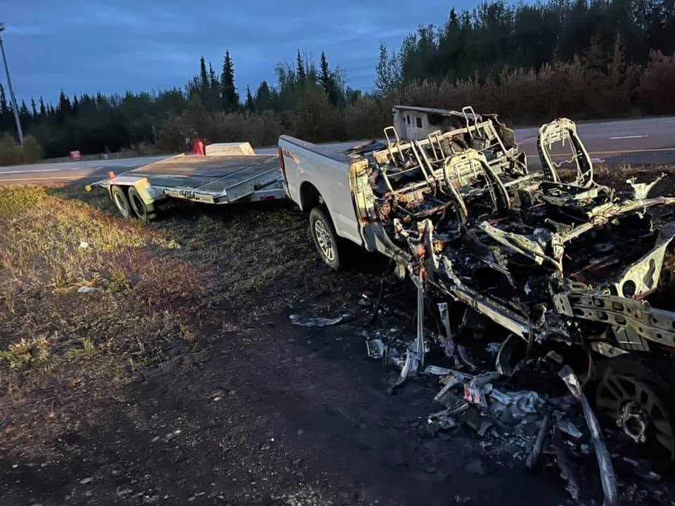 Burned-out tow truck and trailer on a roadside at dusk, with charred wreckage and smoke damage.