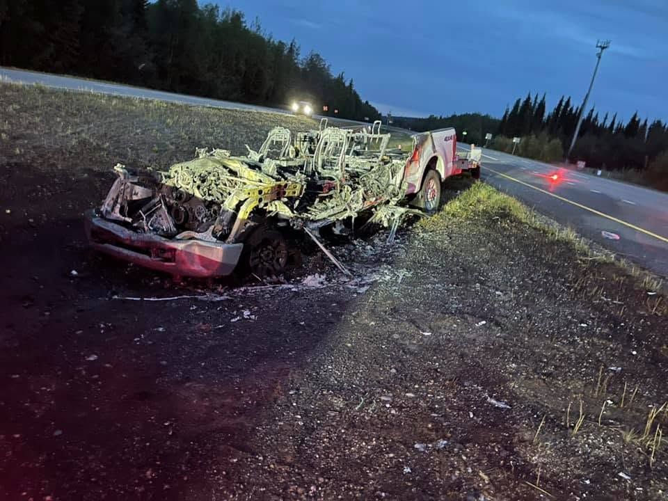 Severely damaged vehicle wreck on a highway shoulder at dusk, with debris and emergency lights nearby