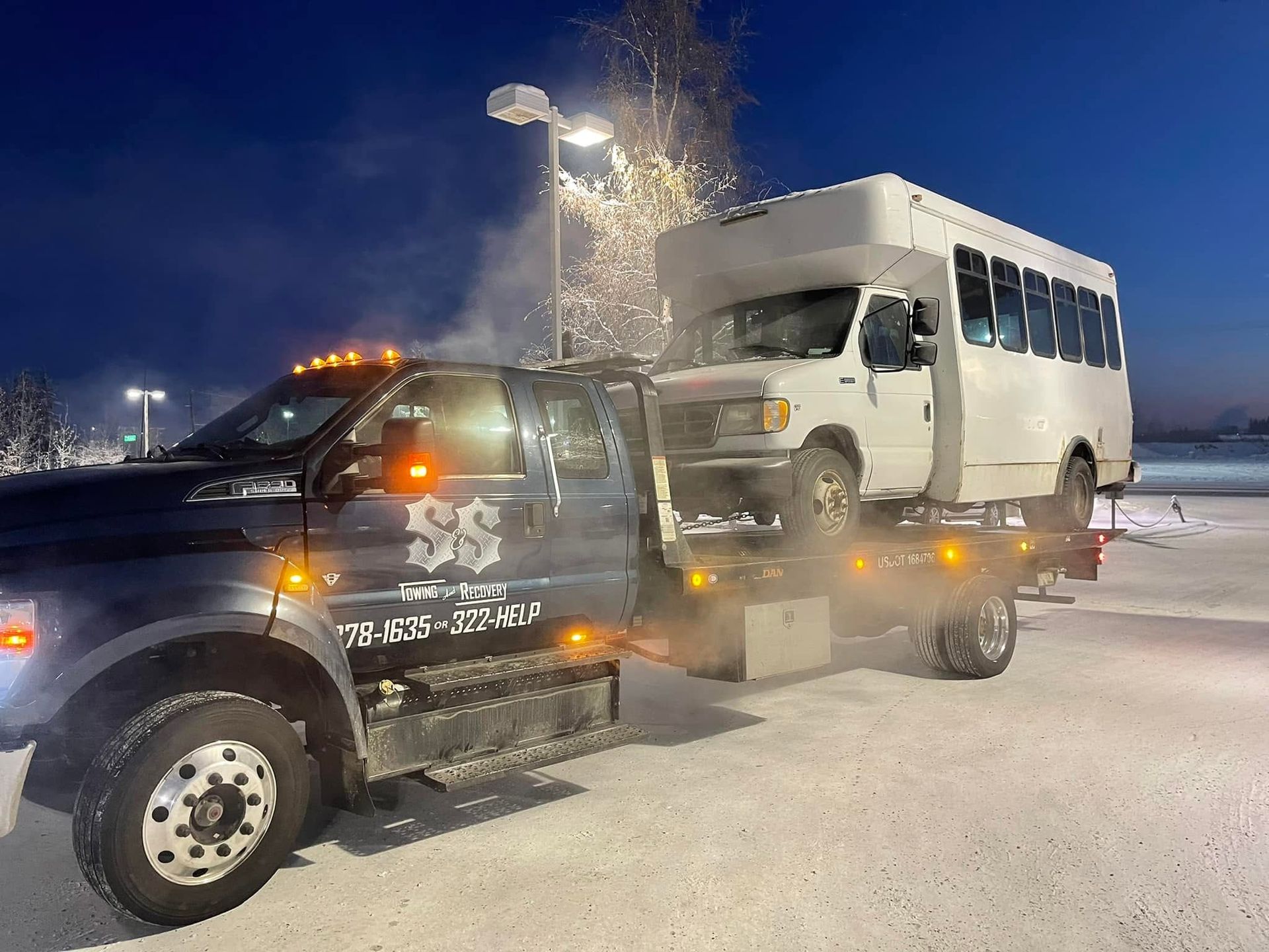 Tow truck hauling a white shuttle bus in a snowy parking lot at night.