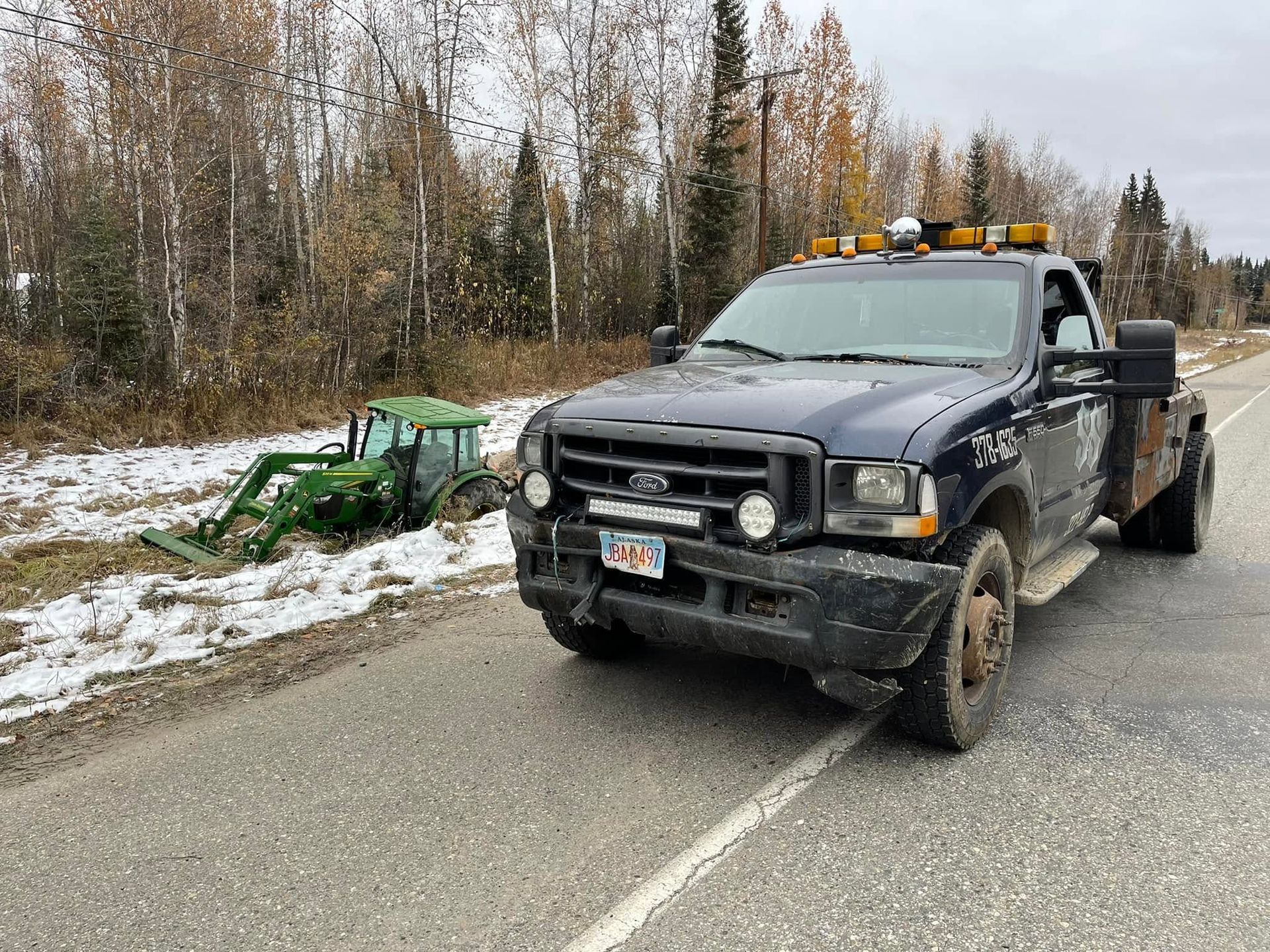 Black tow truck parked on a snowy roadside beside a green snowblower, with trees in the background