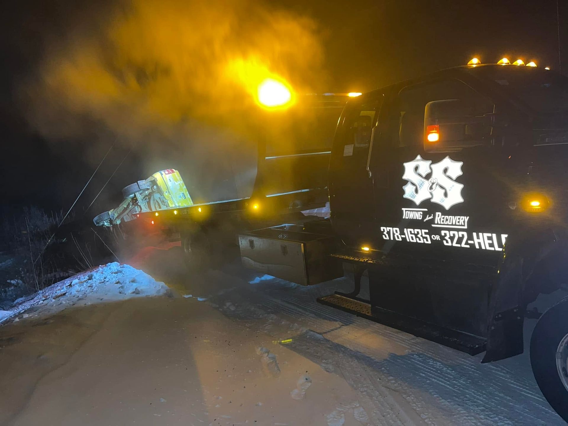 Tow truck with flashing lights at night beside a crashed car on a snowy roadside